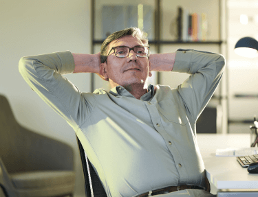 A middle-aged man relaxes in his chair with his hands behind his head.