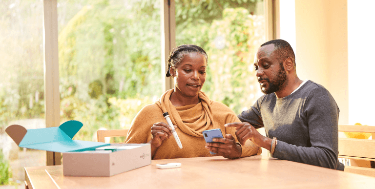 A middle aged man and women sit in conversation at their kitchen table. The woman has the Tempo Pen and Tempo Smart Button in her right hand, the man is pointing at the screen of the smartphone in her left hand