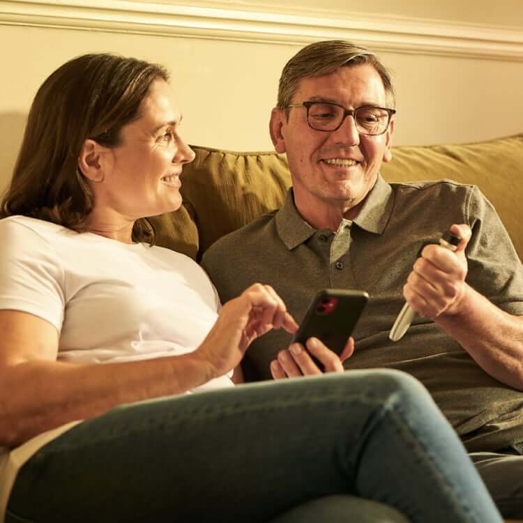 A middle-aged man and woman sit on their couch; the woman holds a smartphone while the man looks at a Tempo Pen with Tempo Smart Button attached.